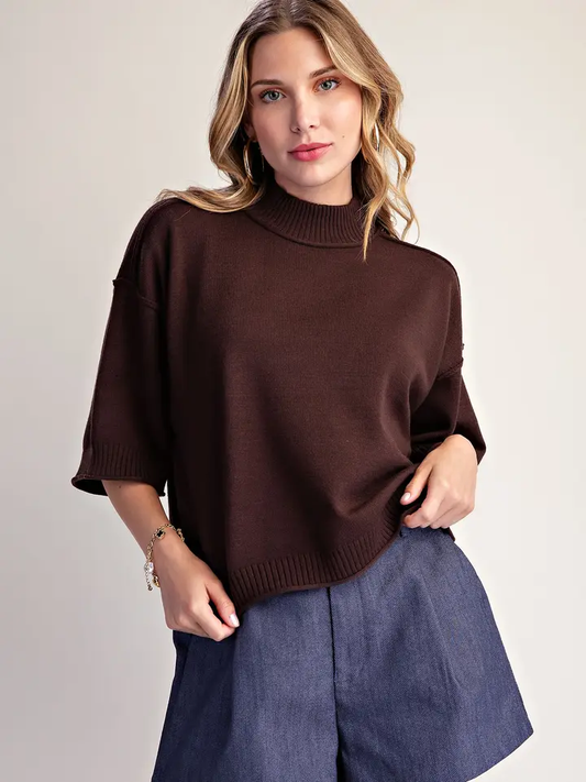 Model wearing a dark brown mock neck sweater with three-quarter sleeves and ribbed trim, styled with a denim skirt