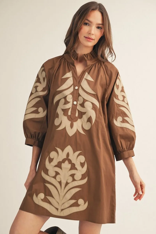 Model wearing a brown cotton shift dress with contrasting floral appliqué, three-quarter sleeves, a ruffled high neckline, and a button-up front